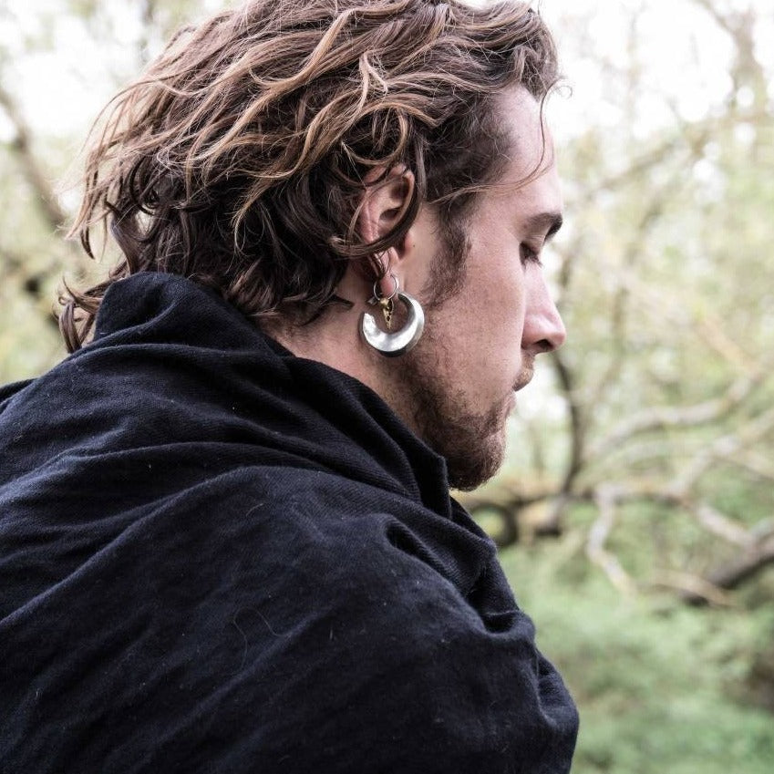 A person with wavy hair and a large crescent-shaped earring is wearing a dark cloak in an outdoor setting. The individual, showcasing stretched ear lobes adorned with HOLO Hoop Dangle Ear Weights in Gold by Kaya Movement (6 gauge), is facing away, looking to the side, against a blurry backdrop of trees and foliage.